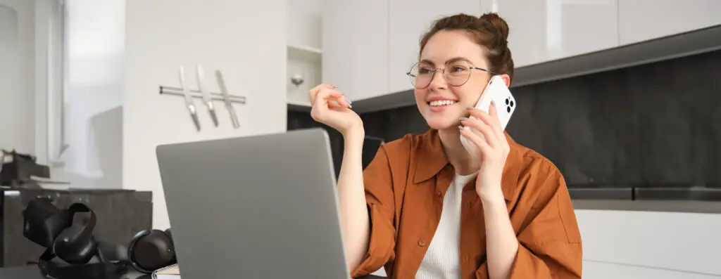 Portrait of young self-employed woman, entrepreneur working from home, freelancer calling client. Girl making an order, talking to someone on phone, sitting in kitchen with laptop